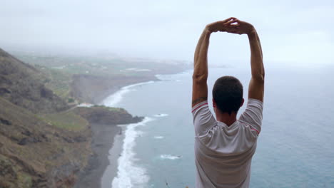 Atop-a-cliff,-a-man-raises-his-hands,-inhaling-the-ocean-breeze-as-he-engages-in-yoga,-embracing-the-soothing-atmosphere